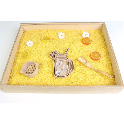 Houten speelbak gevuld met gele speelrijst, met bloemen en citroenschijven. Zeefje en schepje met lange steel, vultray of vulvorm limonadeglas van PLA (geperst tarwestro)